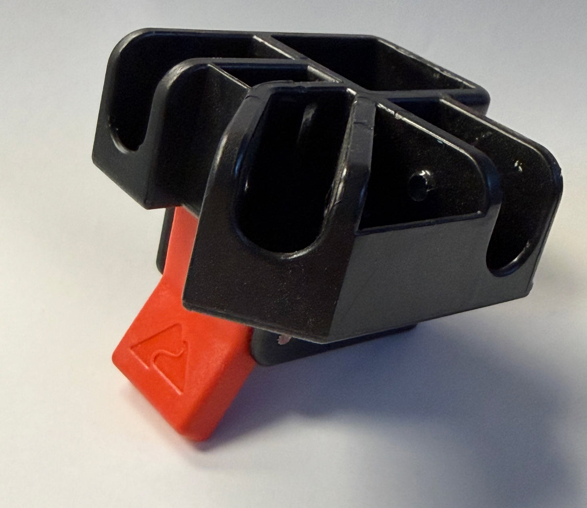 A black plastic multi-groove holder or clip with a bright red triangular push-button switch on the front, photographed on a neutral light gray background.