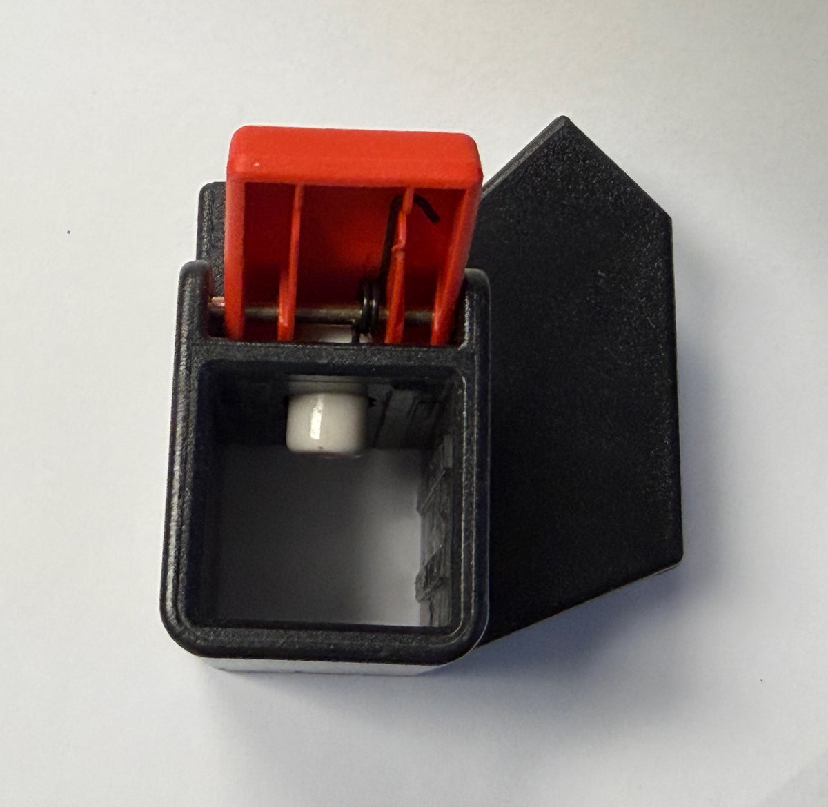 A close-up of a small mechanical switch or toggle component on a plain light background. The product has a black rectangular housing, a red top lever, and a white cylindrical element inside.